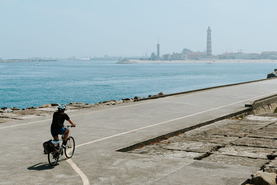 Person cycling along a coastal path with lighthouse and cityscape in view.